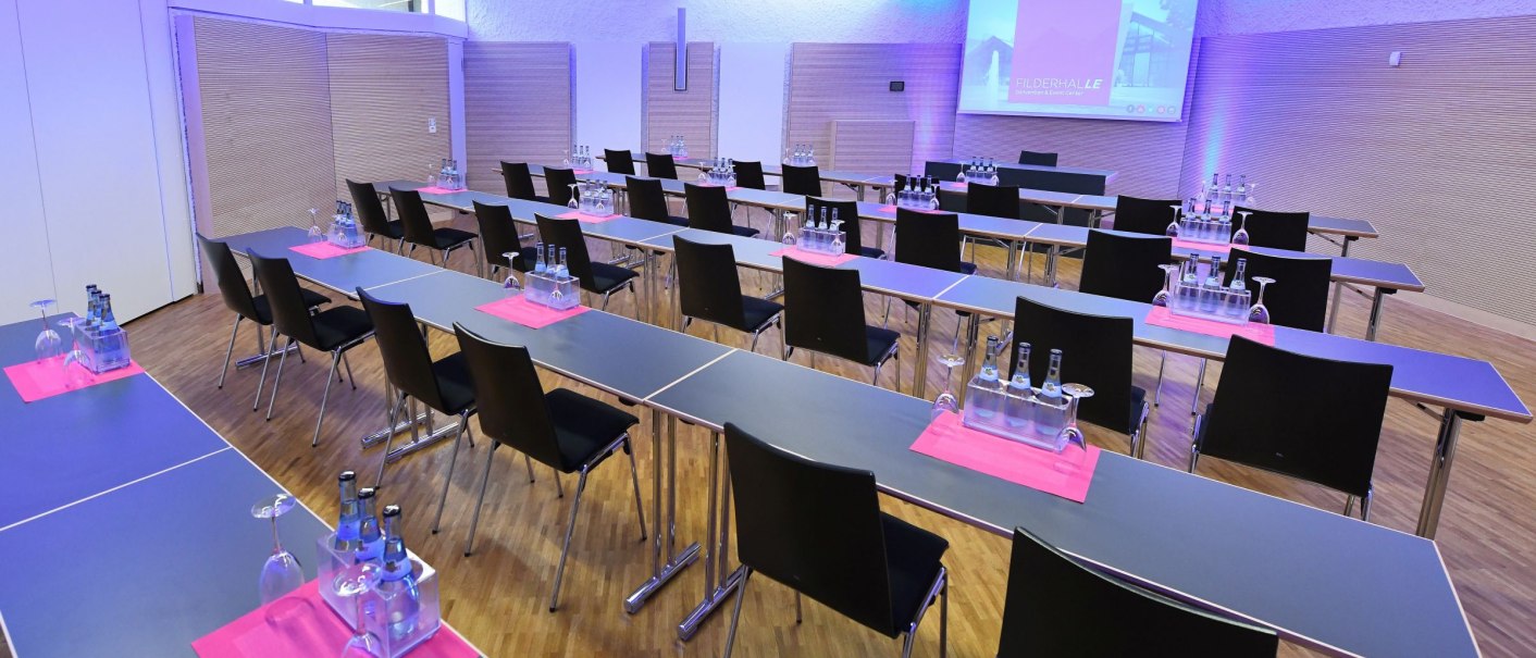 Ein moderner Konferenzraum mit Tischen und Stühlen, ausgestattet mit Wasserflaschen und Gläsern auf pinken Unterlagen. Ein Projektor zeigt 'Filderhalle'., © Guenter E. Bergmann - Photography