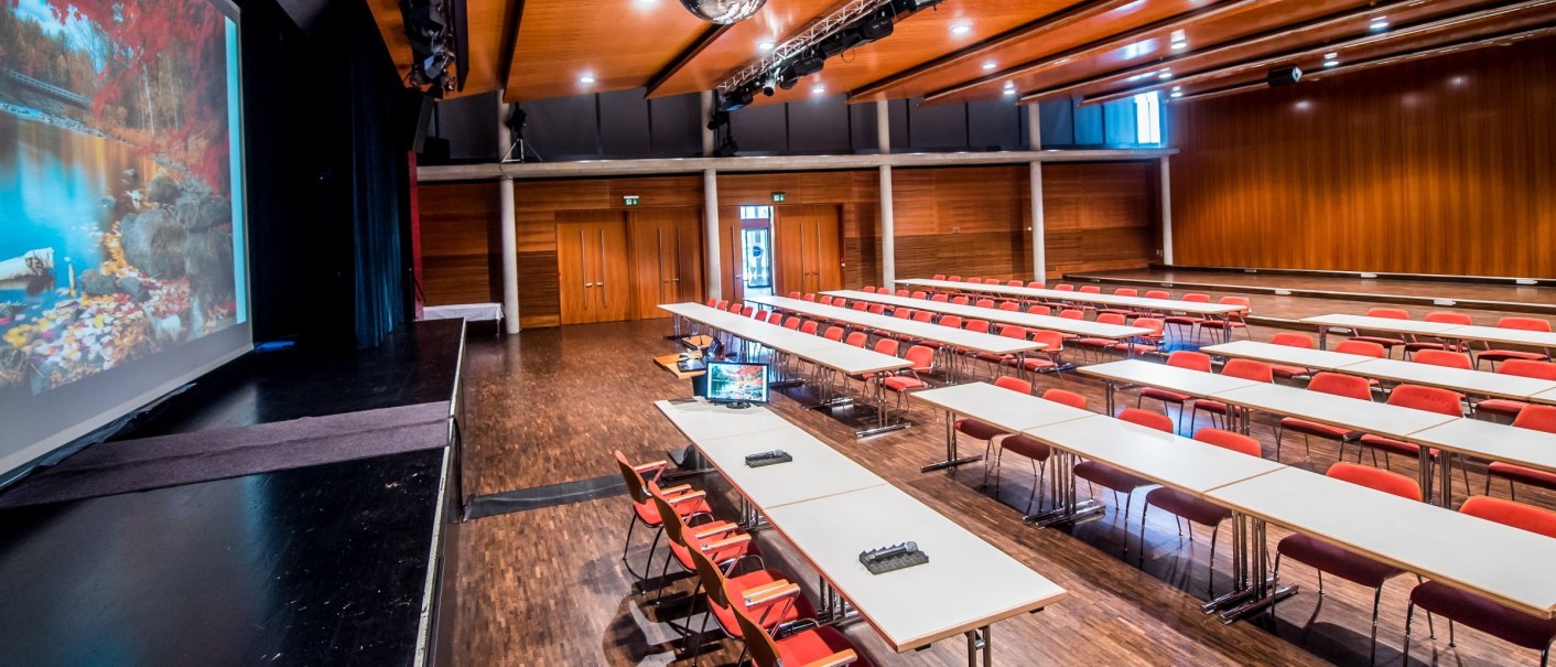 Großer Tagungsraum mit langen Tischen und roten Stühlen, einer Bühne mit Leinwand und einer Discokugel an der Decke., © GIACINTO_CARLUCCI