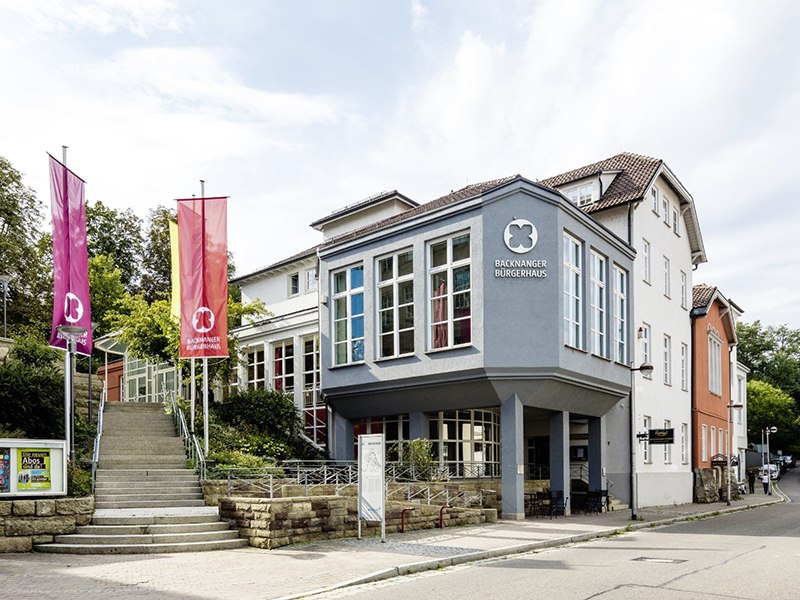 The Backnang community center with a modern façade, large windows and two colorful flags. A staircase leads to the entrance., © © die arge lola, Kai Loges + Andreas Langen The Backnang community center with a modern façade, large windows and two colorful flags. A staircase leads to the entrance., © © die arge lola, Kai Loges + Andreas Langen