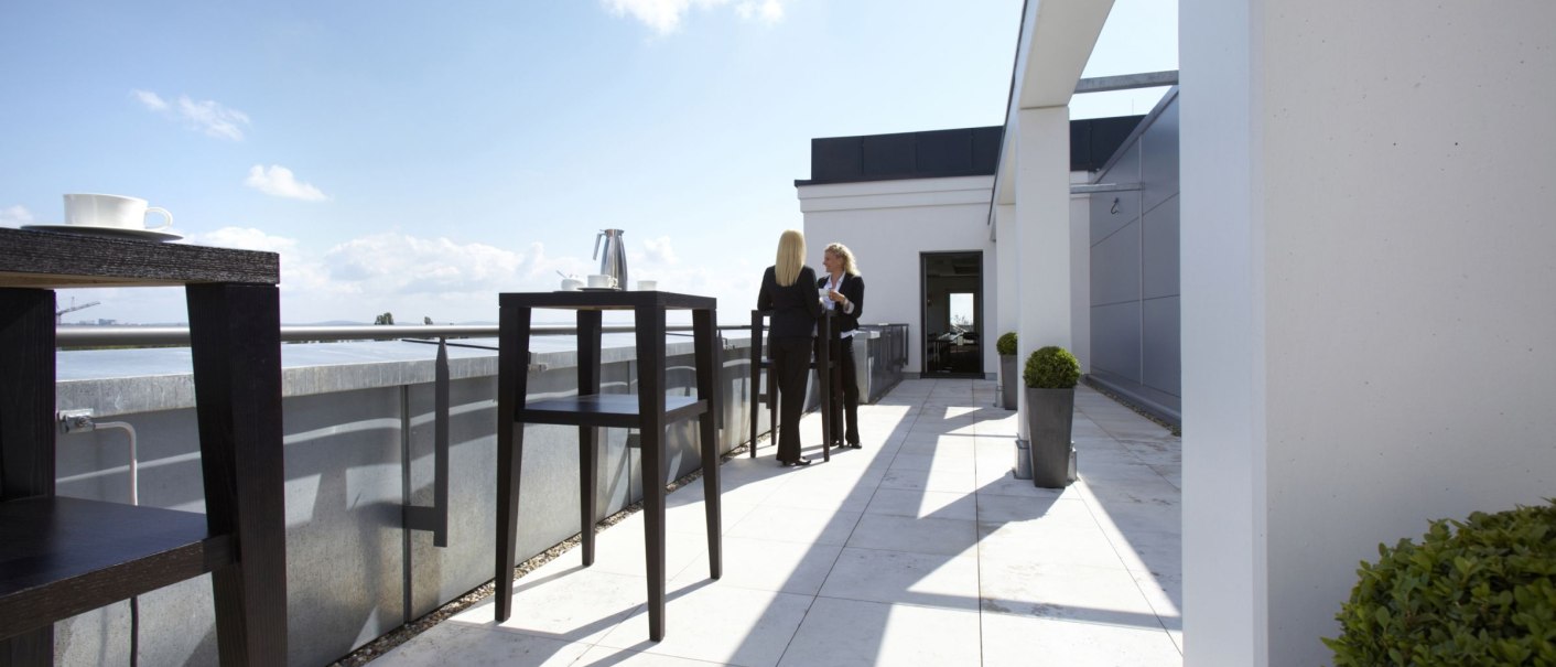 Dachterrasse mit zwei Personen im Gespräch, umgeben von hohen Tischen und Pflanzen. Der Himmel ist blau mit wenigen Wolken., © Parkhotel Stuttgart Messe-Airport GmbH & Co KG Dachterrasse mit zwei Personen im Gespräch, umgeben von hohen Tischen und Pflanzen. Der Himmel ist blau mit wenigen Wolken., © Parkhotel Stuttgart Messe-Airport GmbH & Co KG