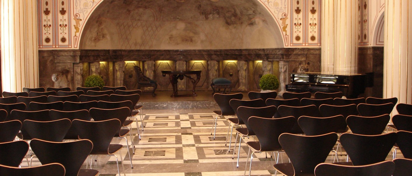 Eleganter Marmorsaal mit Reihen schwarzer Stühle, verzierten Säulen und einem Klavier. Die Wände sind kunstvoll dekoriert., © marmorsaalgmbh