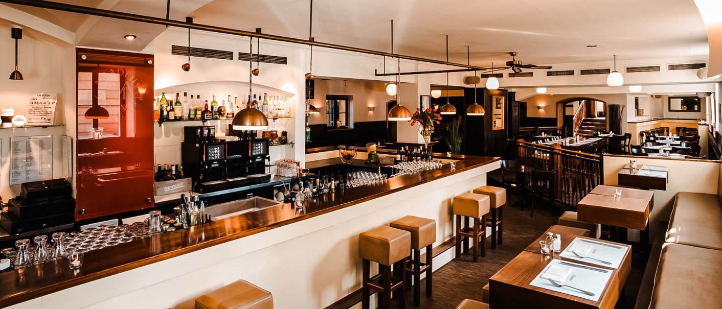 Ein stilvolles Restaurant mit Bar, Holzmöbeln und moderner Beleuchtung. Die Bar ist gut bestückt, und es gibt Sitzgelegenheiten mit gedeckten Tischen., © Amadeus