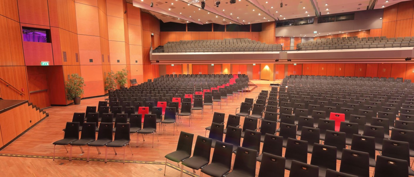 The Europasaal of the Kongresshalle Böblingen with empty rows of chairs, a stage and wooden walls. The ceiling is equipped with lights and technology., © ccbs-gf