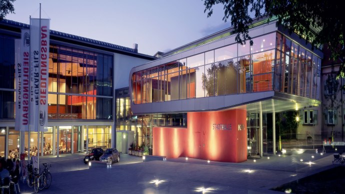 Die Stadthalle K3N bei Dämmerung, moderne Glasarchitektur mit beleuchtetem Vorplatz und Fahnenmasten., © k3nmaisch