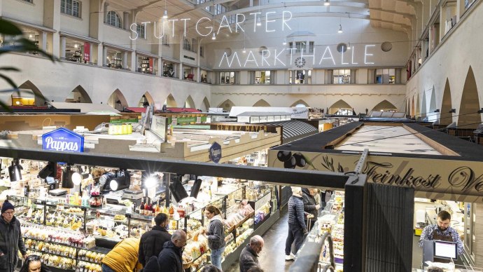 Die Stuttgarter Markthalle mit beleuchteten Ständen und vielen Besuchern. Ein großes Schild mit 'Stuttgarter Markthalle' hängt von der Decke., © SMG, Sarah Schmid