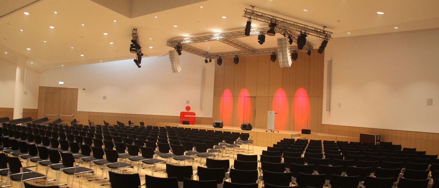Moderner Konferenzsaal mit leeren Stuhlreihen, Bühne und Beleuchtung. Ein rotes Sparkassen-Logo ist an der Wand sichtbar., © ccbs