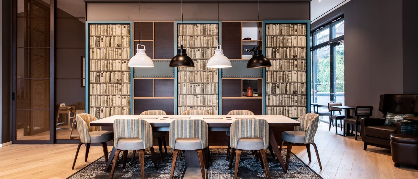 Moderner Besprechungsraum mit großem Tisch, gepolsterten Stühlen und Hängelampen. Bücherregale als Wanddekoration, Holzboden und große Fenster., © Guenter Lenz Moderner Besprechungsraum mit großem Tisch, gepolsterten Stühlen und Hängelampen. Bücherregale als Wanddekoration, Holzboden und große Fenster., © Guenter Lenz