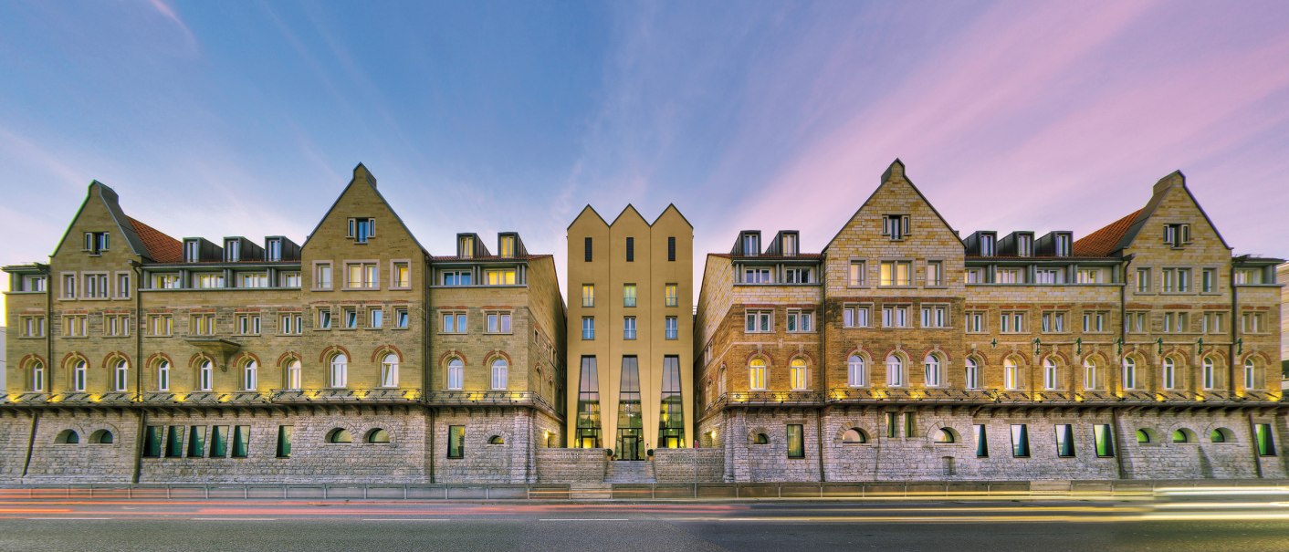 Das Arcotel Camino zeigt eine symmetrische Fassade aus Stein mit modernen Elementen, beleuchtet bei Sonnenuntergang. Eine Straße verläuft davor., © ARCOTEL Hotels