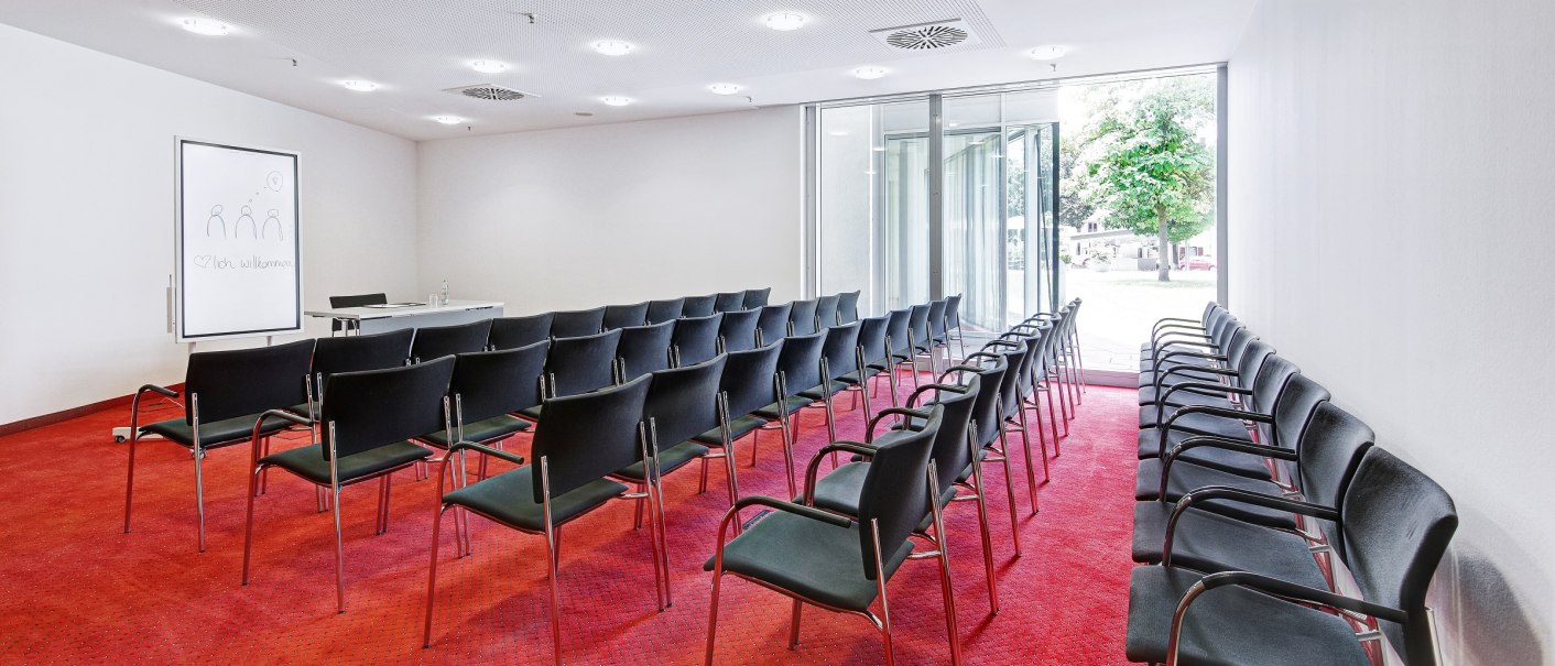 A conference room with red carpeting, rows of black chairs and a flipchart. Large windows offer a view of the outside., © Martin Frey A conference room with red carpeting, rows of black chairs and a flipchart. Large windows offer a view of the outside., © Martin Frey