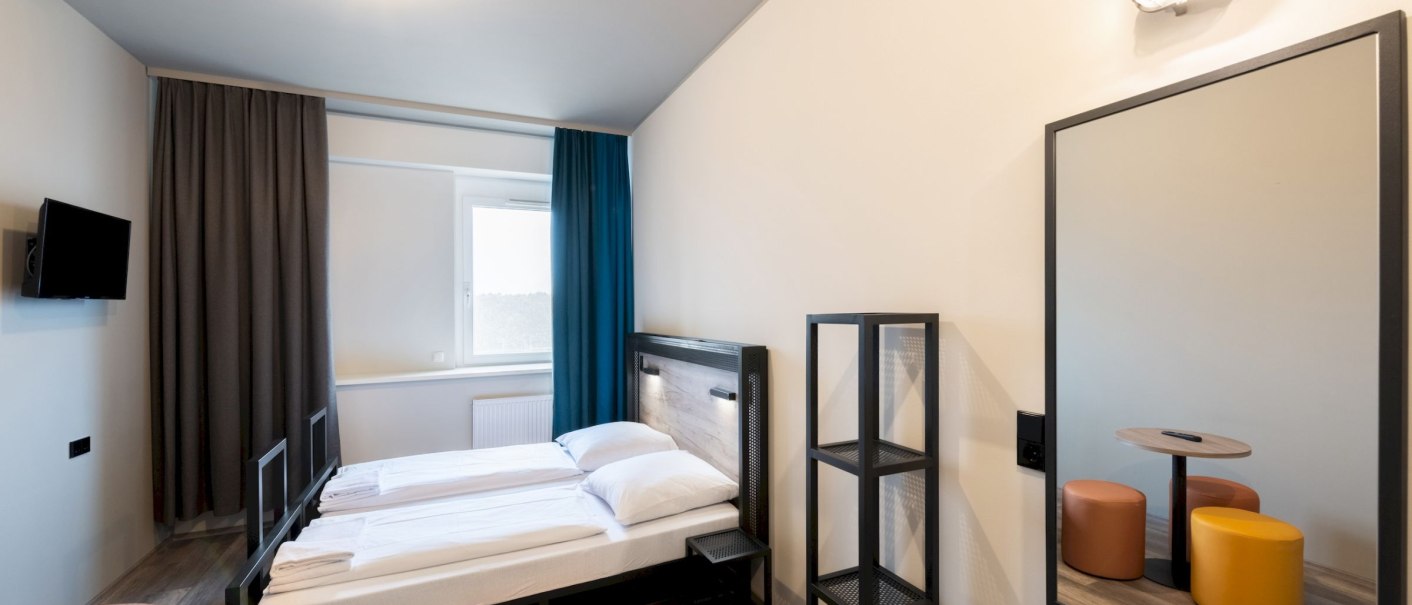 Modernes Hotelzimmer mit zwei Einzelbetten, kleinem Tisch, Hockern, Fernseher und Regal. Helle Wände, Holzboden und Vorhänge in Grau und Blau., © Jan Engelhardt