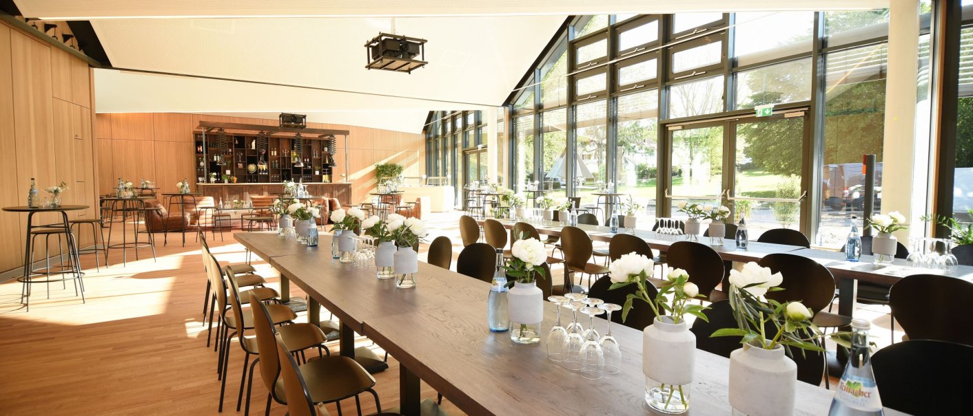 Moderner Veranstaltungsraum mit langen Tischen, Stühlen und großen Fenstern. Elegante Dekoration mit Blumenvasen und Gläsern auf den Tischen., © Guenter E. Bergmann - Photography