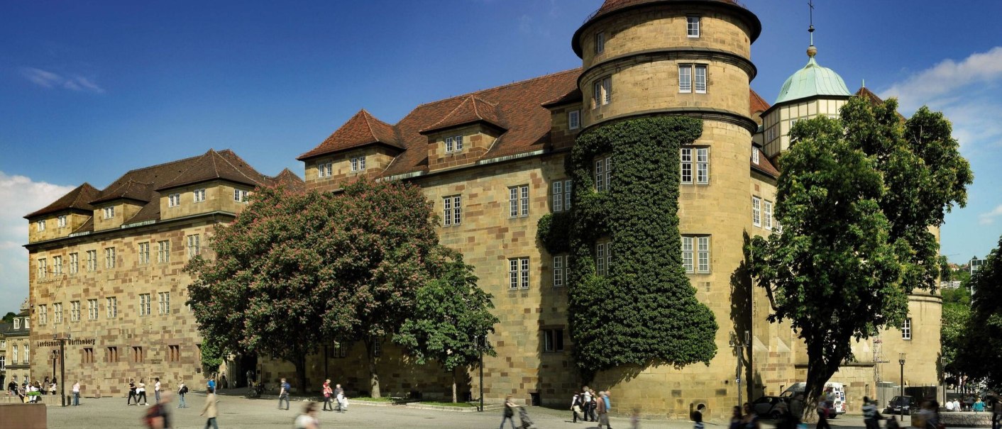 Das Alte Schloss in Stuttgart mit bewachsenen Türmen und Bäumen im Vordergrund. Menschen spazieren auf dem Platz bei sonnigem Wetter., © Stuttgart-Marketing GmbH Florian Selig