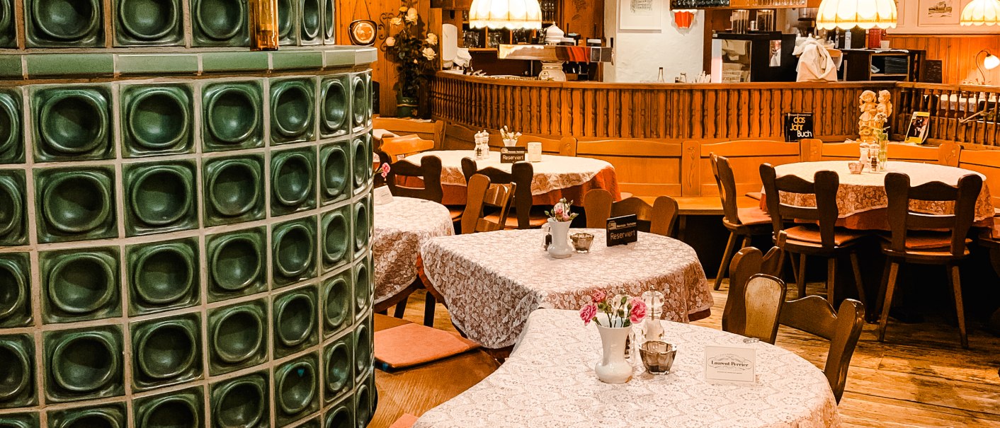 Cozy restaurant with wooden furniture, tablecloths and a green tiled stove. Lamps hanging from the ceiling, bar in the background., © Weinstube Kachelofen