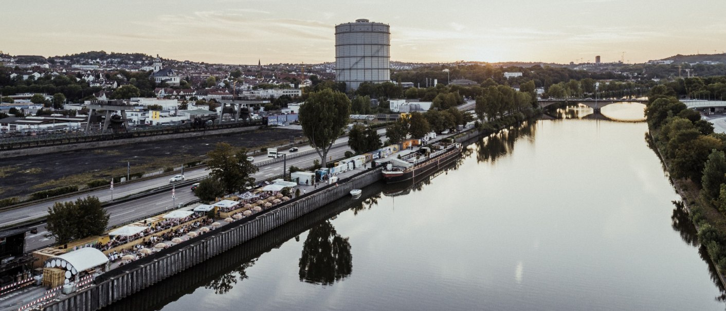Aerial view of a river with promenade, trees and a large round building in the background at sunset., &copy; Fridas Pier