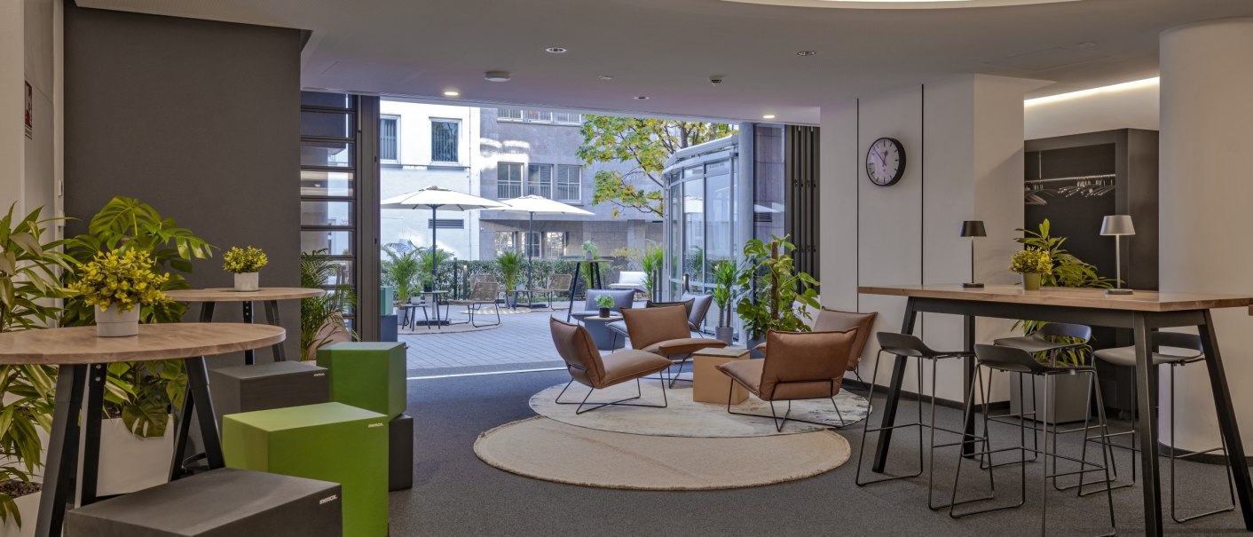 Modern room with high tables, stools and plants. A terrace with parasols in the background. Bright, inviting atmosphere., © R&D Dürr