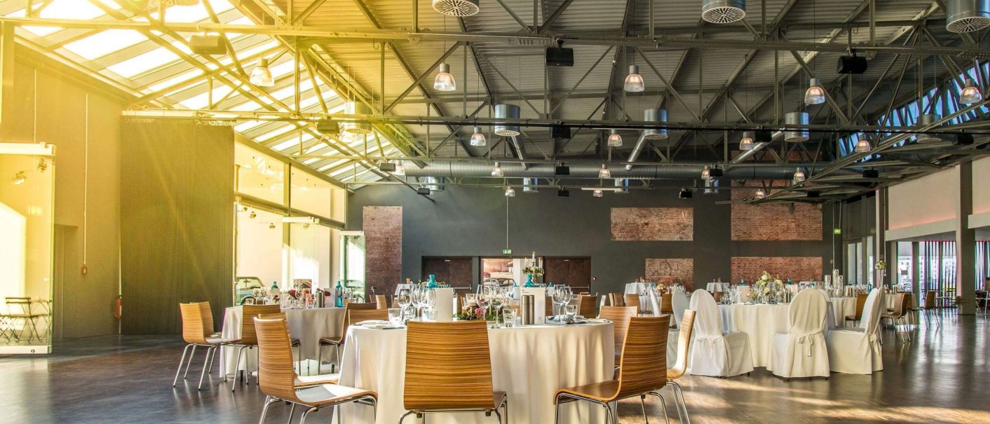 Large hall with industrial design, covered tables and chairs. Sunlight streams through the windows, creating a warm atmosphere., © rauschenbergercatering