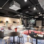 Moderne Lounge mit schwarzen Tischen, hellen Hockern und Regalen. Ein Fahrrad hängt an der Wand, Pflanzen und bunte Teppiche dekorieren den Raum., © Design Offices GmbH