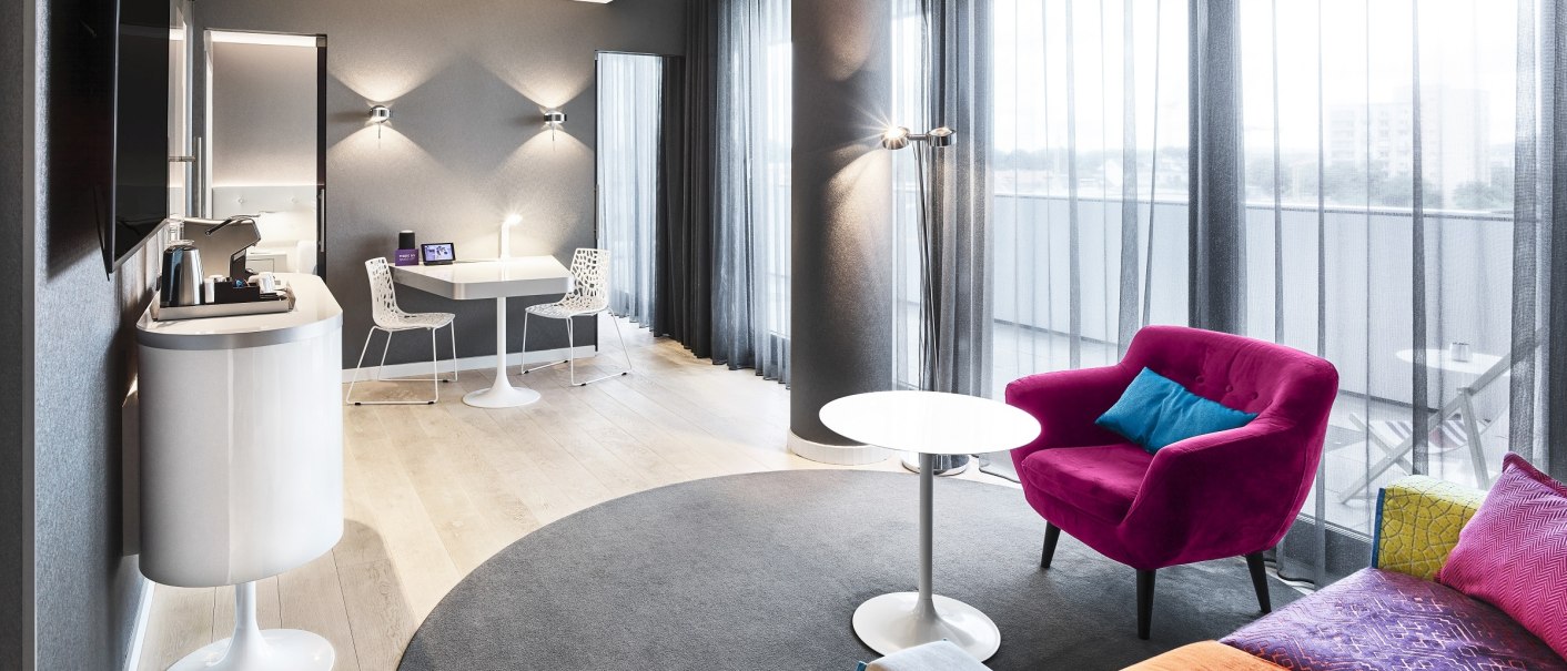 Modern hotel room with colorful armchair, sofa, table and large windows. Light wooden floors and stylish furnishings create an inviting atmosphere., &copy; FOTOGRAFIE FREI