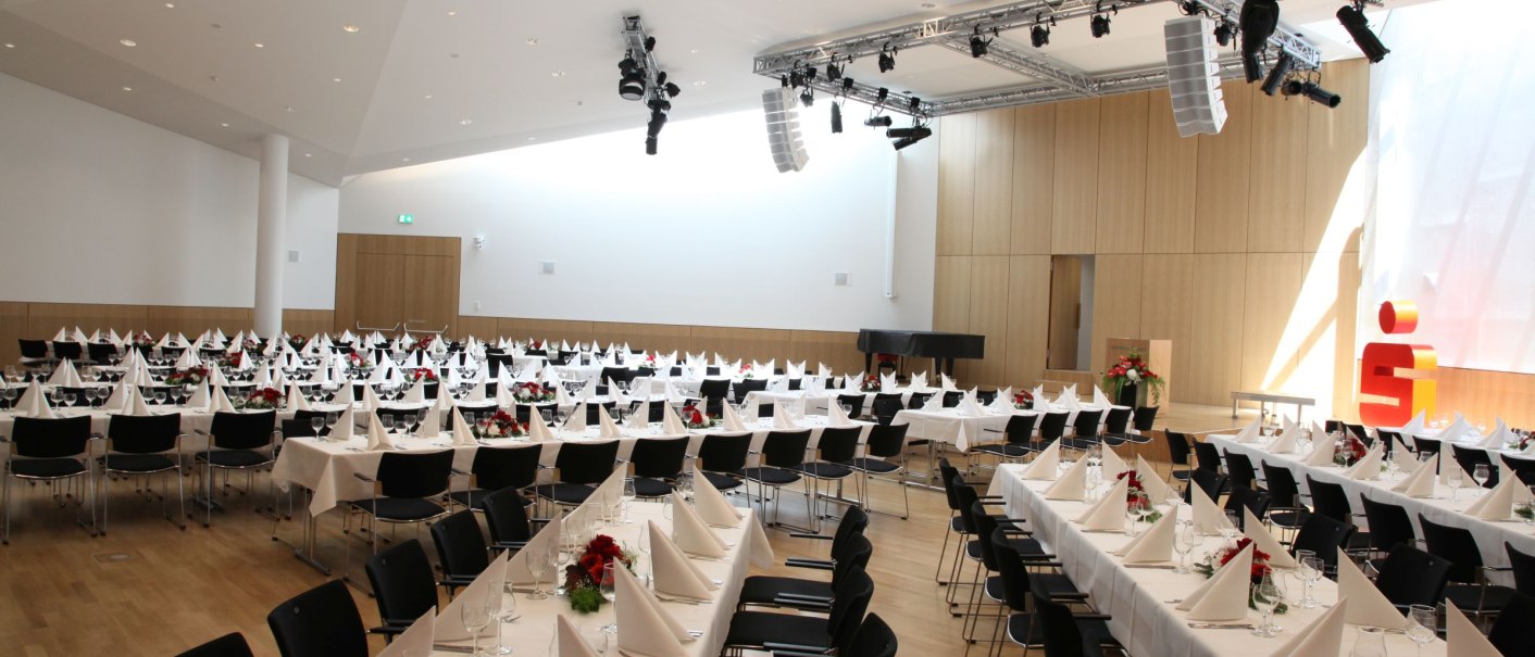 Large function room with set tables and chairs, ready for an event. Bright, modern furnishings with stage and lighting., © ccbs
