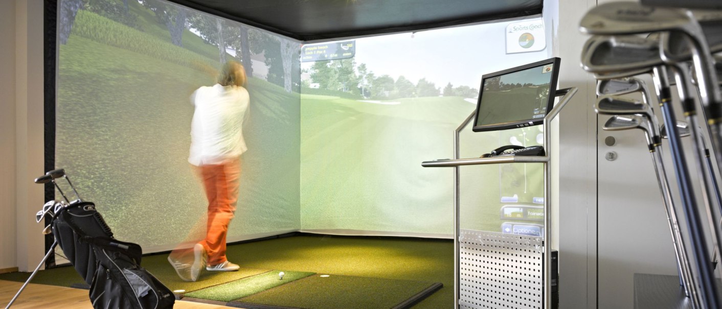 Person spielt in einer Indoor-Golfanlage mit Simulator. Ein Bildschirm zeigt das Spiel, Golfschläger stehen bereit., © Parkhotel Stuttgart Messe-Airport GmbH & Co KG Person spielt in einer Indoor-Golfanlage mit Simulator. Ein Bildschirm zeigt das Spiel, Golfschläger stehen bereit., © Parkhotel Stuttgart Messe-Airport GmbH & Co KG