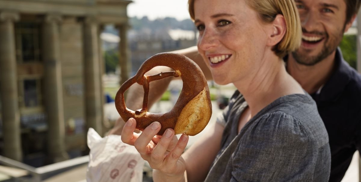 Frau hält eine schwäbische Brezel, lächelnder Mann im Hintergrund, Gebäude im Hintergrund., © TMBW, Christoph Düpper