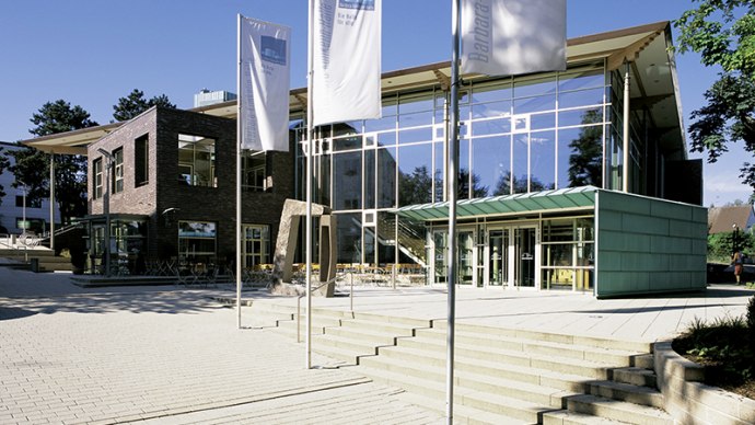 Modern hall with glass fa&ccedil;ade and flags in the foreground, surrounded by trees and blue sky., &copy; Barbara-K&uuml;nkelin-Halle