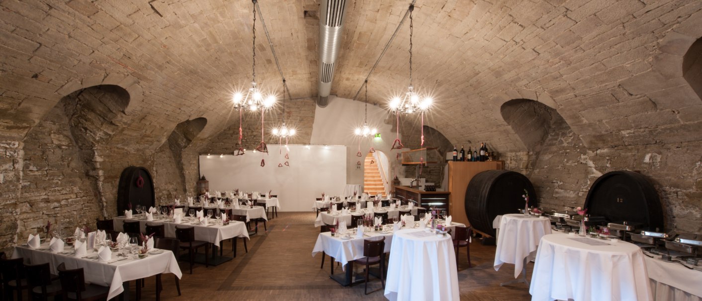 Ein eleganter Weinkeller mit gedeckten Tischen, Kronleuchtern und großen Weinfässern an den Wänden. Der Raum ist für ein Event vorbereitet., © Landschloss Korntal Ein eleganter Weinkeller mit gedeckten Tischen, Kronleuchtern und großen Weinfässern an den Wänden. Der Raum ist für ein Event vorbereitet., © Landschloss Korntal