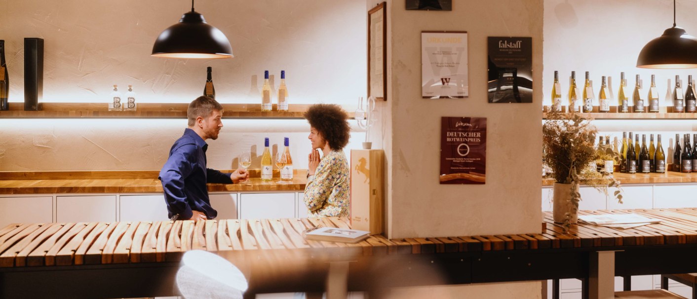 Zwei Personen unterhalten sich in einer Vinothek. Weinflaschen stehen auf Regalen, Urkunden an der Wand. Gemütliche Atmosphäre mit Holztischen., © SMG, Alwin Maigler
