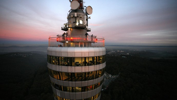 Ein Fernsehturm mit beleuchteter Aussichtsplattform bei Sonnenuntergang. Der Himmel ist rosa gef&auml;rbt, und die Landschaft erstreckt sich im Hintergrund., &copy; SWR Media Services GmbH / Achim Mende