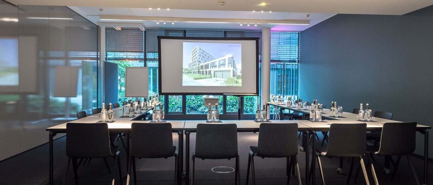 Modern conference room with U-shaped tables, chairs and a large projector screen. Water bottles and notepads are distributed on the tables., © © Marc Gilardone Photographer