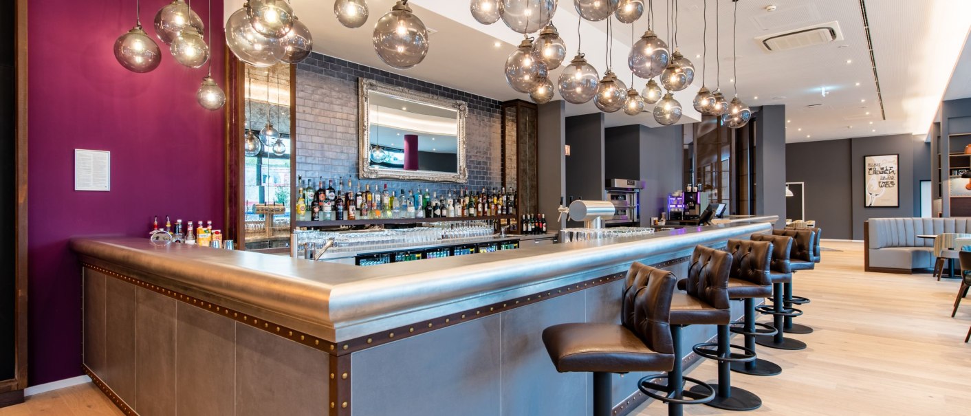 Moderne Bar mit Metalltheke, braunen Hockern und hängenden Glaskugellampen. Im Hintergrund sind Flaschen und ein Spiegel zu sehen., © Guenter Lenz