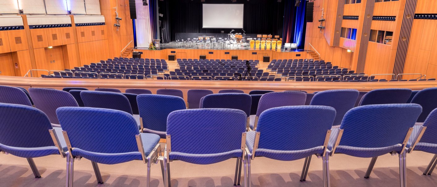Gro&szlig;er Saal der Stadthalle Sindelfingen mit leeren blauen St&uuml;hlen und B&uuml;hne. Holzw&auml;nde und Beleuchtung schaffen eine warme Atmosph&auml;re., &copy; ccbs