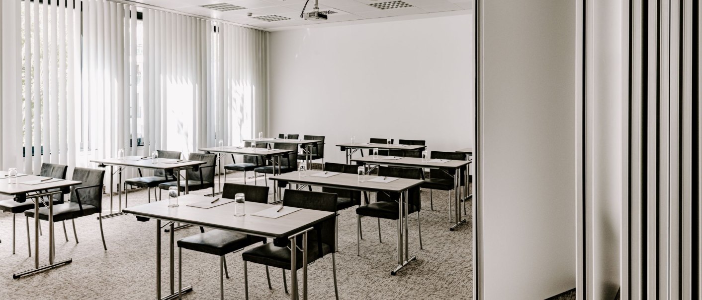 Ein moderner Tagungsraum mit Tischen und Stühlen, einem Deckenprojektor und großen Fenstern mit Jalousien. Der Raum ist hell und funktional eingerichtet., © VHE Mo.Stuttgart