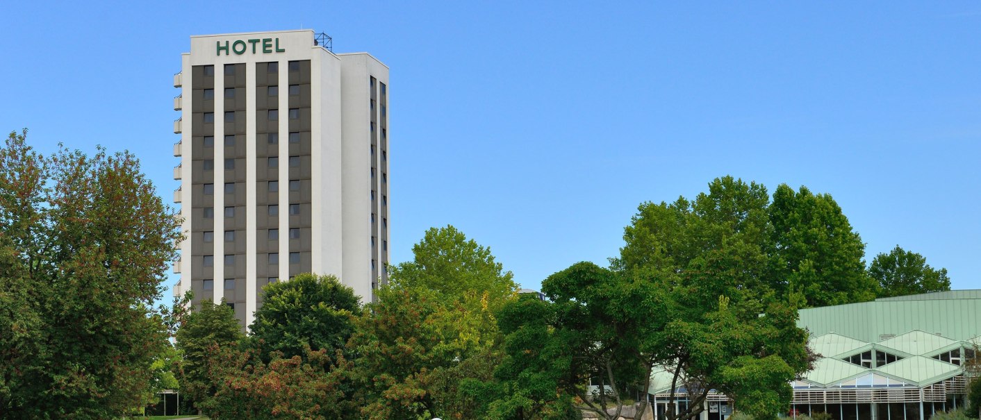 A tall hotel building with the inscription 'HOTEL' stands behind a green meadow and trees under a clear blue sky., &copy; DH STUDIO Dirk Holst