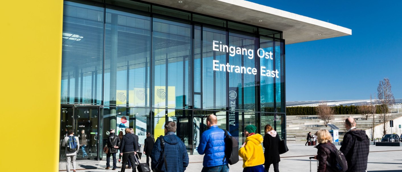 People walk to the East Entrance of Messe Stuttgart. Modern glass fa&ccedil;ade, blue sky, some carrying luggage., &copy; Landesmesse Stuttgart / Valentin Marquardt