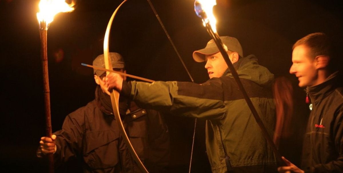 Drei Männer schießen nachts mit Bögen, beleuchtet von Fackeln. Einer spannt den Bogen, während die anderen zuschauen. Dunkle Umgebung., © Teamgeist Süd GmbH Drei Männer schießen nachts mit Bögen, beleuchtet von Fackeln. Einer spannt den Bogen, während die anderen zuschauen. Dunkle Umgebung., © Teamgeist Süd GmbH
