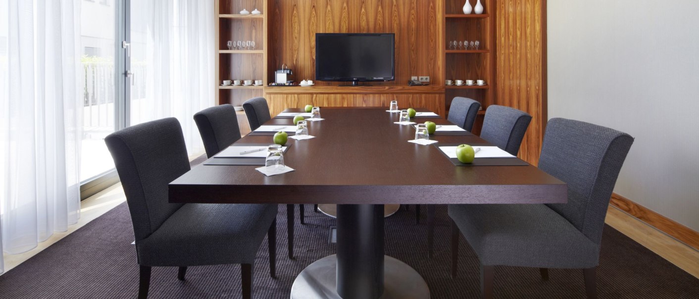 Konferenzraum mit großem Tisch, Stühlen, Notizblöcken und grünen Äpfeln. Im Hintergrund ein Fernseher und Regale mit Geschirr., © Parkhotel Stuttgart Messe-Airport GmbH & Co KG Konferenzraum mit großem Tisch, Stühlen, Notizblöcken und grünen Äpfeln. Im Hintergrund ein Fernseher und Regale mit Geschirr., © Parkhotel Stuttgart Messe-Airport GmbH & Co KG