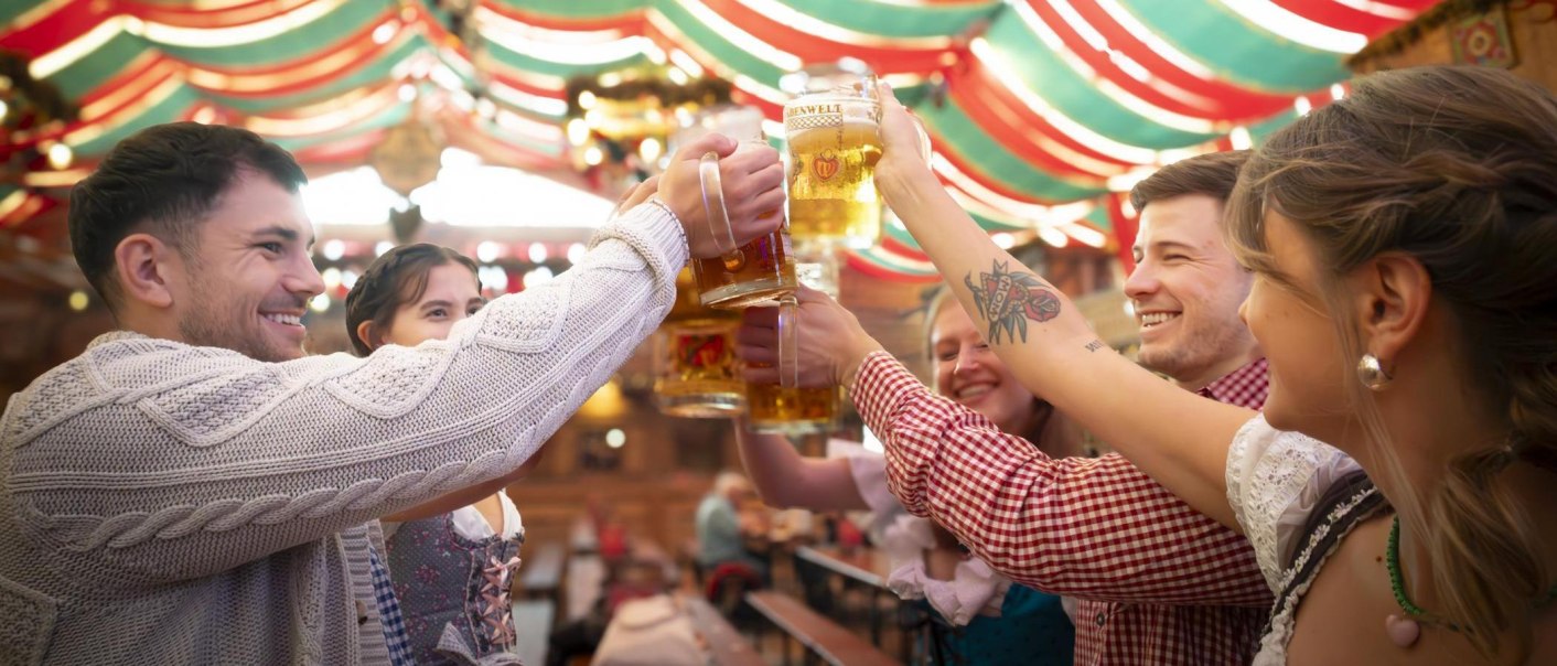 Menschen in traditioneller Kleidung sto&szlig;en mit Bierkr&uuml;gen in einem festlich dekorierten Zelt an. Fr&ouml;hliche Stimmung auf einem Volksfest., &copy; Stuttgart-Marketing GmbH, Sarah Schmid