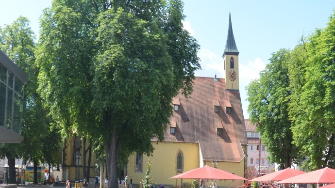 The Kreuzkirche N&uuml;rtingen church with its striking tower, surrounded by green trees and red parasols on a sunny square., &copy; kkmaisch_gf