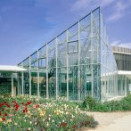 Modernes Glasgebäude der Stadthalle Sindelfingen, umgeben von bunten Blumenbeeten unter blauem Himmel., © ccbs