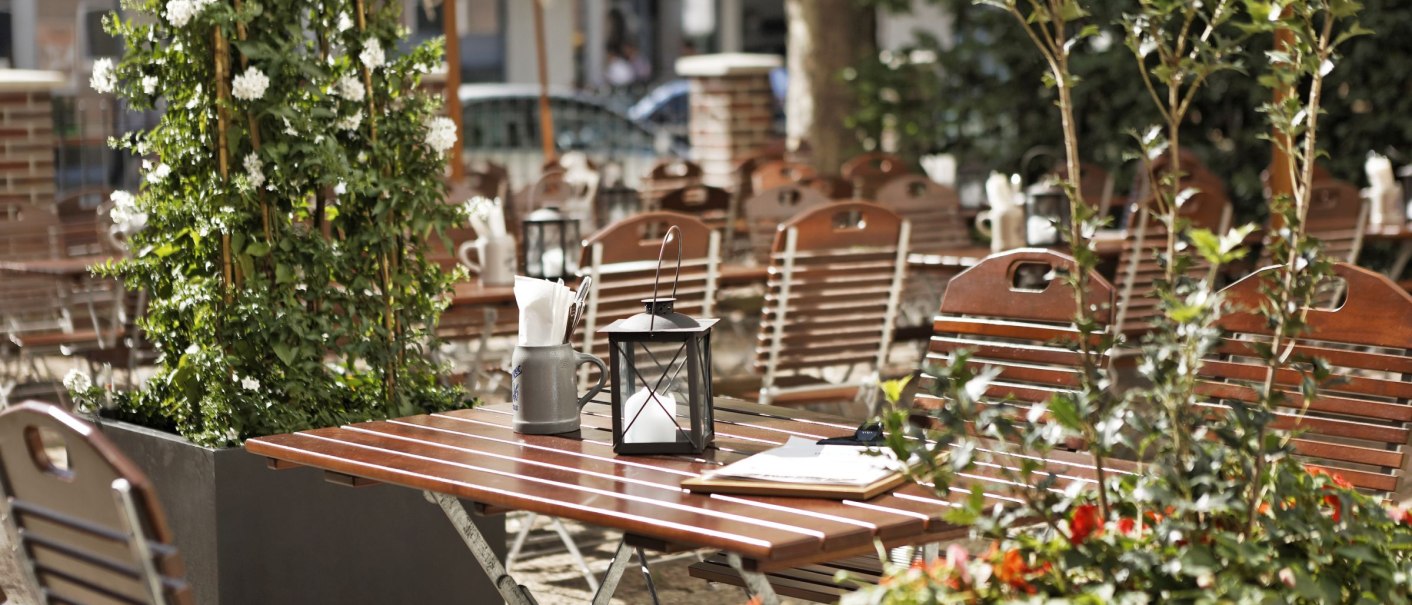 Ein leerer Biergarten mit Holztischen und -stühlen, umgeben von grünen Pflanzen und bunten Blumen. Im Hintergrund sind Sonnenschirme zu sehen., © Parkhotel Stuttgart Messe-Airport GmbH & Co KG Ein leerer Biergarten mit Holztischen und -stühlen, umgeben von grünen Pflanzen und bunten Blumen. Im Hintergrund sind Sonnenschirme zu sehen., © Parkhotel Stuttgart Messe-Airport GmbH & Co KG