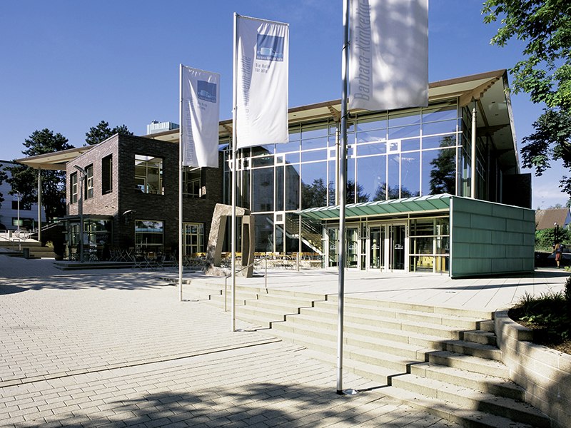 Modern hall with glass fa&ccedil;ade and flags in the foreground, surrounded by trees and blue sky., &copy; Barbara-K&uuml;nkelin-Halle