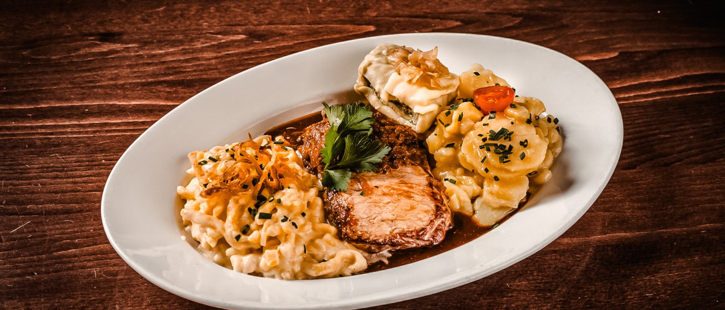 A plate of roast, spaetzle, Maultasche and potato salad, garnished with parsley, served on a wooden table., © Amadeus