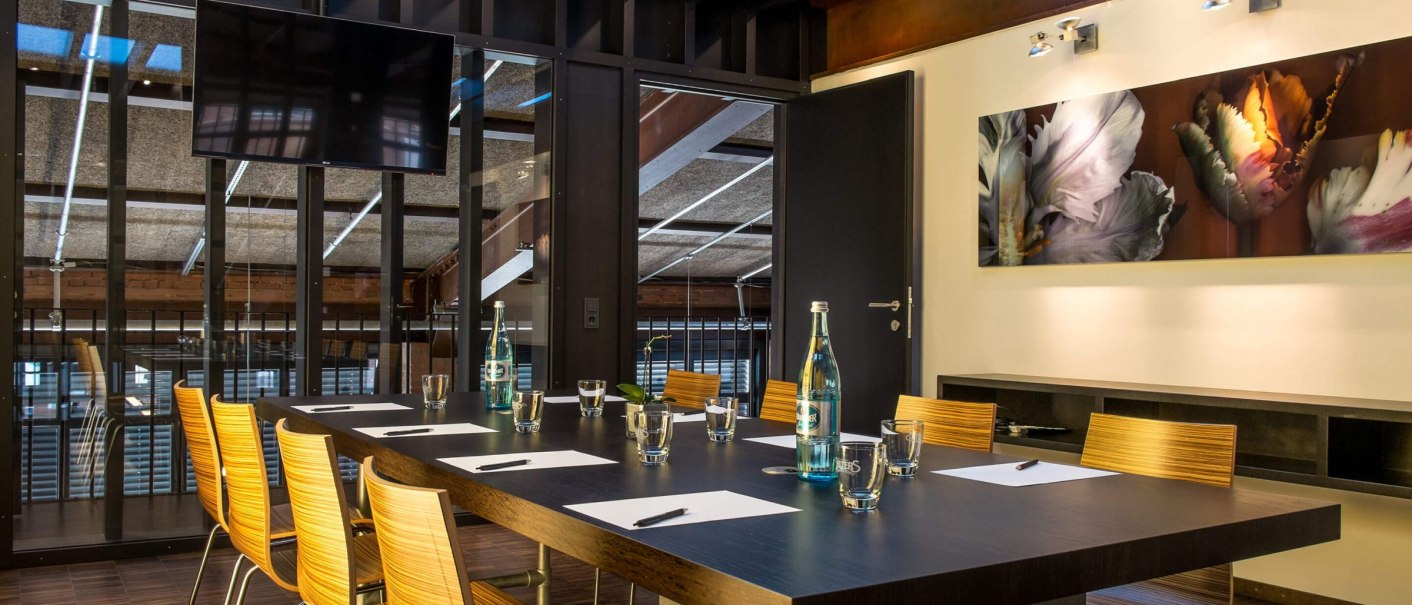 Moderner Meetingraum mit großem Holztisch, Stühlen, einem Fernseher und Kunst an der Wand. Wasserflaschen und Gläser sind auf dem Tisch platziert., © rauschenbergercatering