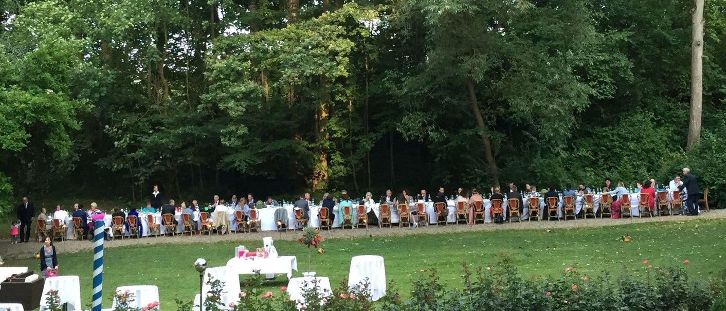 Lange Tafel im Freien, umgeben von Bäumen. Menschen sitzen an festlich gedeckten Tischen im Park von Burg Schaubeck. Sommerliche Atmosphäre., © GFA_GF