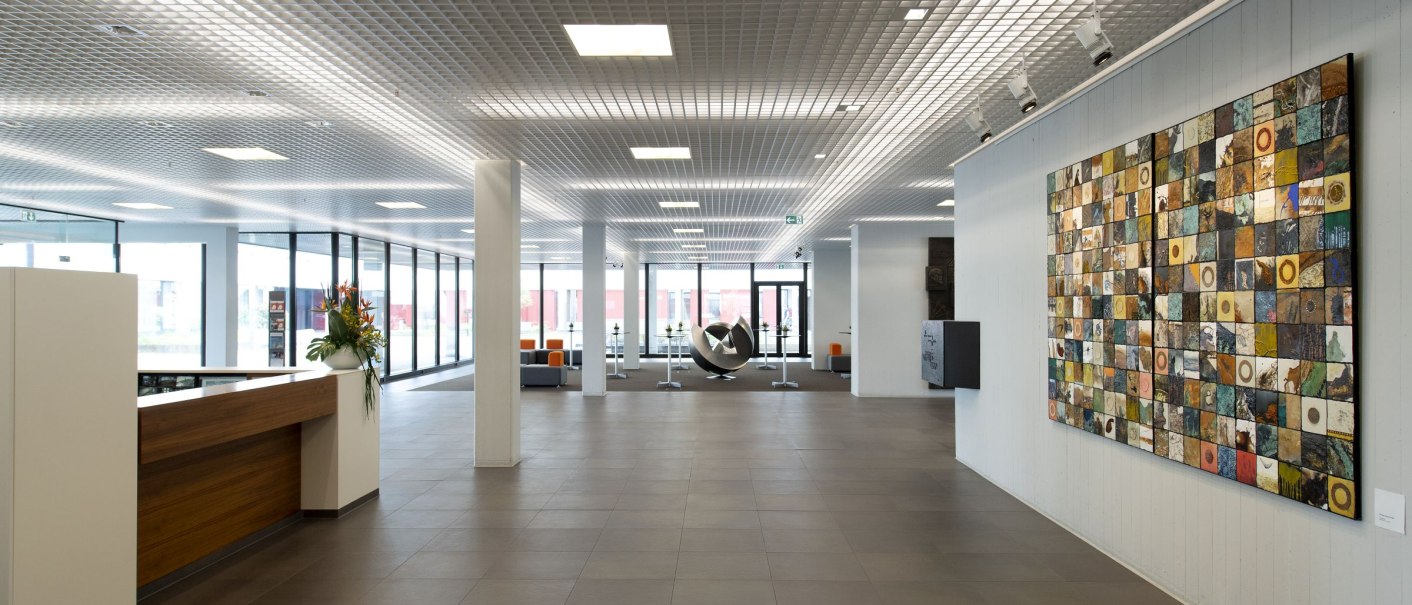 Modernes Foyer mit großem Kunstwerk an der Wand, Empfangstresen links und Skulptur im Hintergrund. Helle, offene Gestaltung mit viel Glas., © Geno-Haus