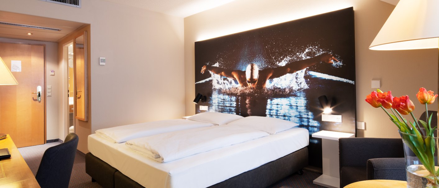 Modernes Hotelzimmer mit Doppelbett, großem Schwimmerbild an der Wand, Schreibtisch und Vase mit Tulpen., © eriksonhotel_gf
