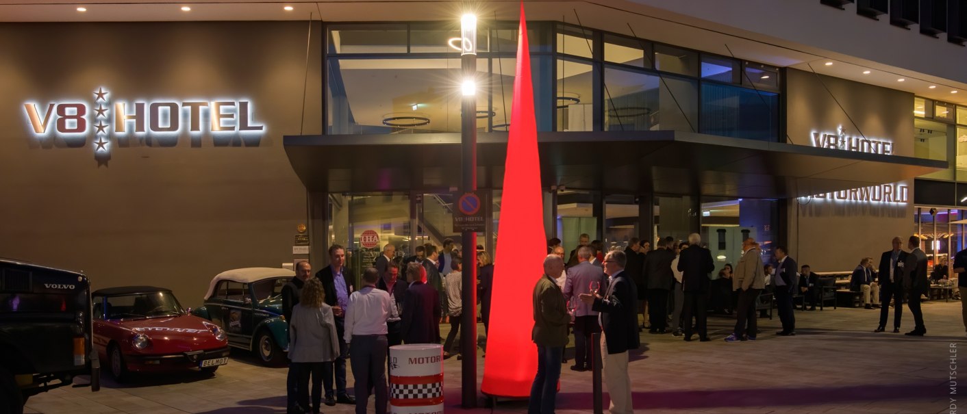 People gather at night in front of the V8 Hotel in the MOTORWORLD Region Stuttgart. Illuminated entrance and classic cars in the foreground., &copy; Hardy Mutschler