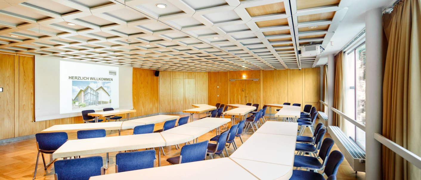 Konferenzraum mit blauen Stühlen, weißen Tischen und Holzwänden. Eine Leinwand zeigt 'Herzlich Willkommen'. Große Fenster mit Vorhängen an der Seite., © Tobias Fröhner Photography