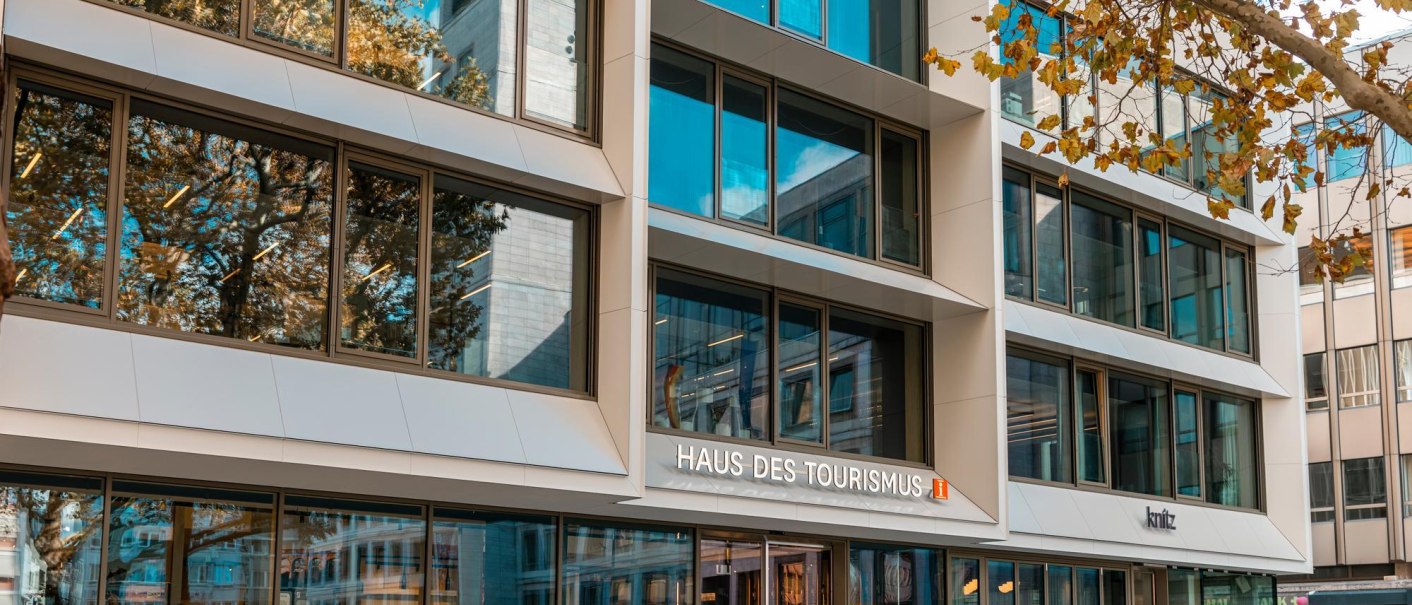 Modern façade of the 'Haus des Tourismus' with large windows and autumnal trees in the foreground. People stand in front of the building., © SMG Stuttgart Marketing GmbH - Sarah Schmid Modern façade of the 'Haus des Tourismus' with large windows and autumnal trees in the foreground. People stand in front of the building., © SMG Stuttgart Marketing GmbH - Sarah Schmid
